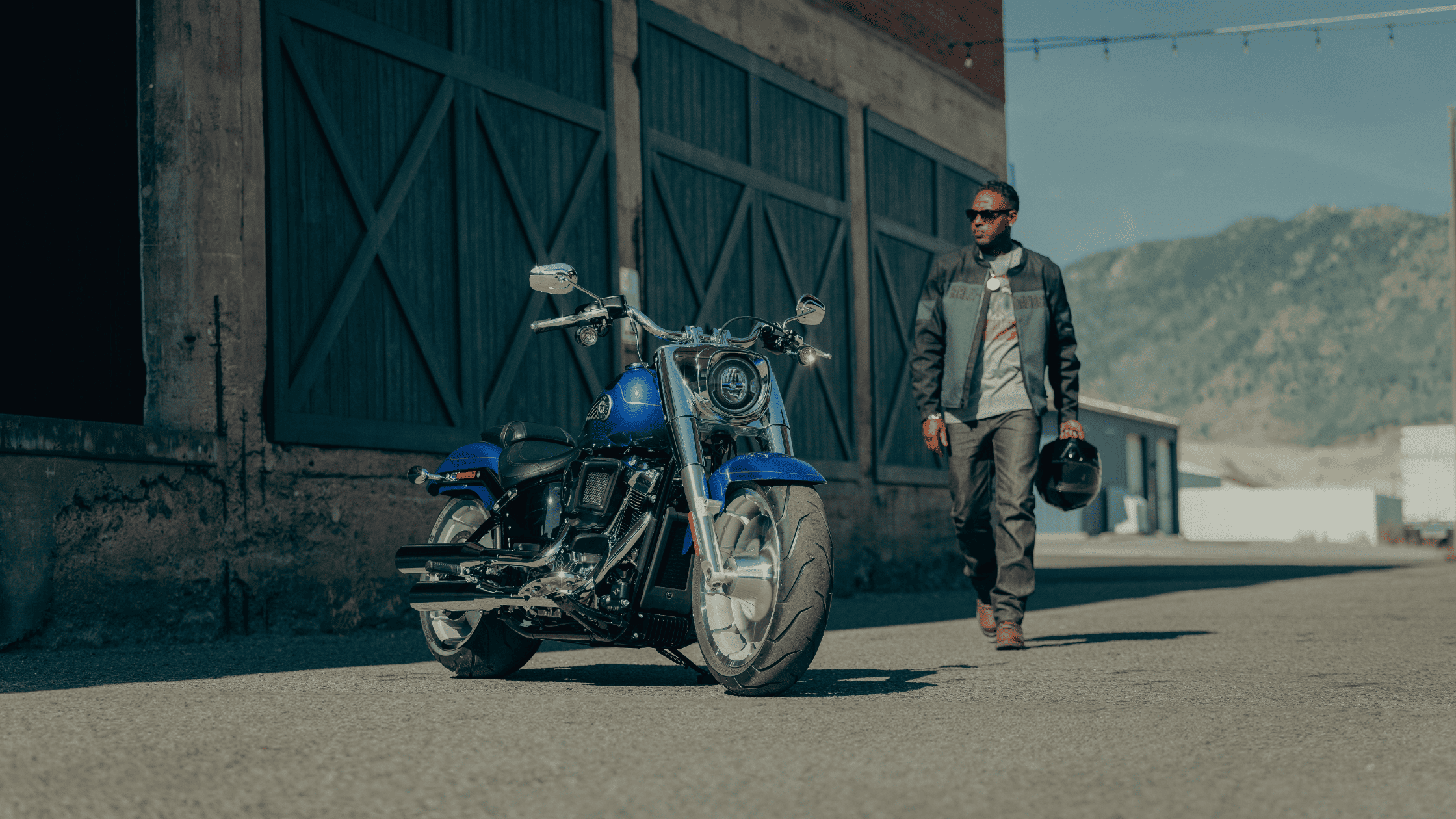 Man walks toward 2025 Harley-Davidson® Fat Boy® parked beside warehouse with mountains in background