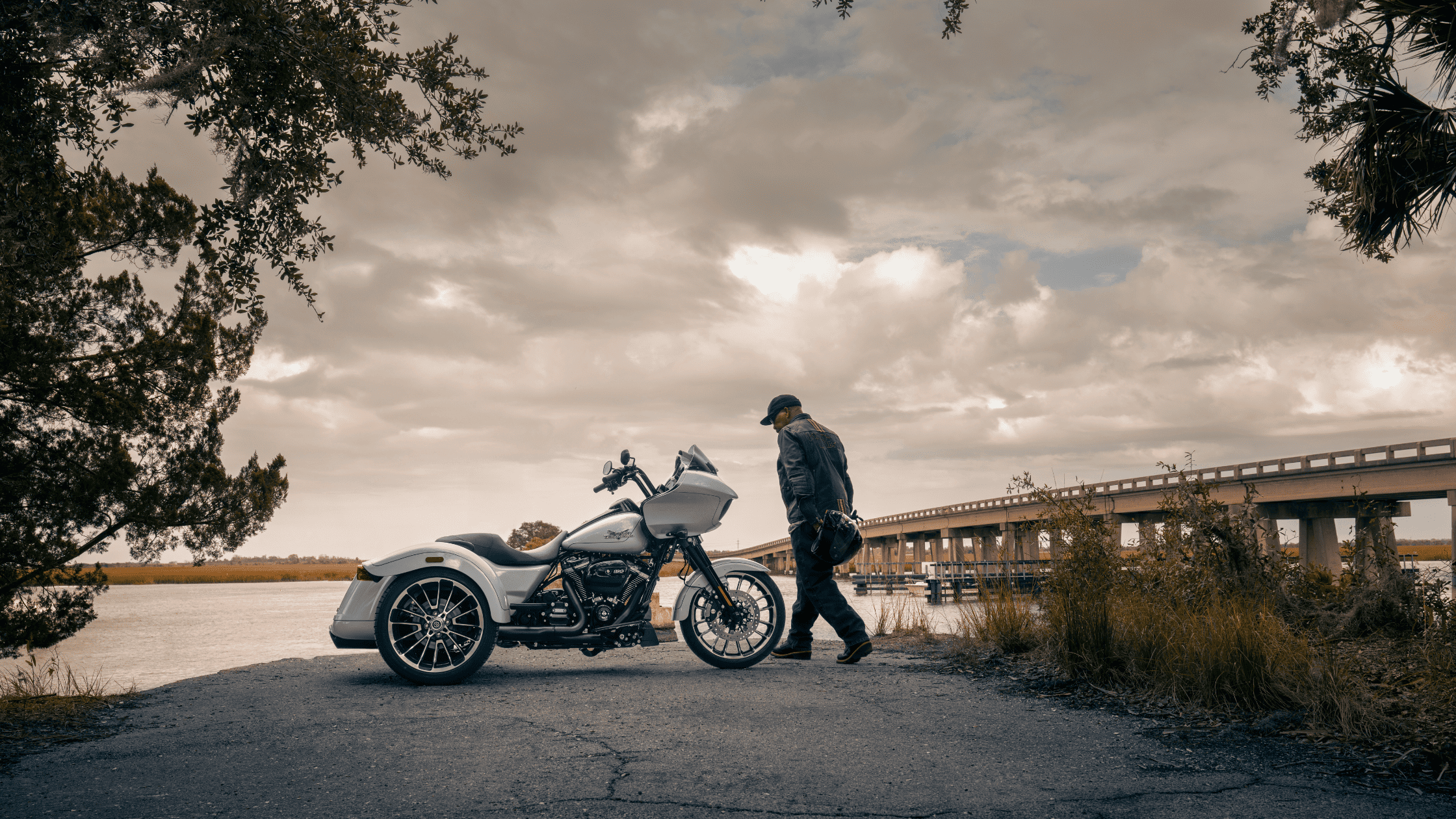 2025 Harley-Davidson® Road Glide® 3 gray trike side profile with rider beside scenic river bridge