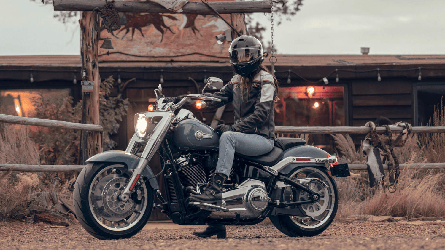 A rider sitting on a 2024 Harley-Davidson&reg; Fat Boy&reg; 114 in front of a rustic cabin, with the motorcy...
